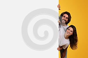 Joyous couple playfully peeking around a blank white space for copy or advertisement