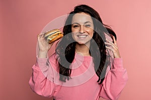 Joyous dark-haired gorgeous young woman posing on the pink background with her favorite nourishing sandwich. Fast food