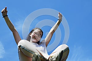 A joyous girl sits and holds her hands up against the blue sky