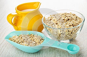 Jug with milk, blue spoon with oat flakes, flakes in bowl on wooden table