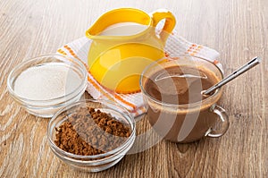 Jug of milk on napkin, bowls with cocoa powder, sugar, cocoa with milk, spoon in cup on wooden table