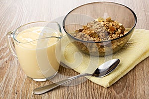 Jug with yogurt, bowl with muesli on napkin, spoon on wooden table