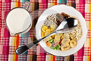 Jug of yogurt with cereals, muesli dried fruits, corn flakes, oatmeal, sunflower seeds, banana chips and spoon in bowl on napkin