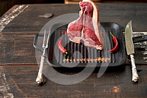 A juicy, aged t-bone steak in a standing position in a grill pan with sprinkled spices and red chili pods and a table knife