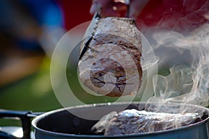 Juicy Angus steak frying in iron cast pan with smoke and Tongs on blurred nature background , cooking party picnic outdoor