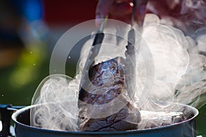 Juicy Angus steak frying in iron cast pan with smoke and Tongs on blurred nature background , cooking party picnic outdoor