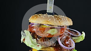 A juicy big burger with a double bun and a double cutlet with greens and vegetables stapled with a knife rotates on a