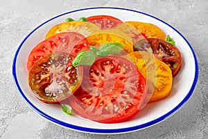 Juicy colorful sliced tomatoes with fresh basil leaves on a plate on a gray concrete background. Healthy summer salad