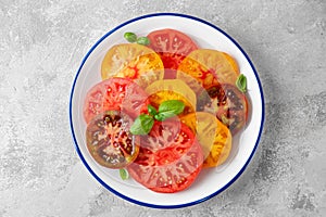 Juicy colorful sliced tomatoes with fresh basil leaves on a plate on a gray concrete background. Healthy summer salad