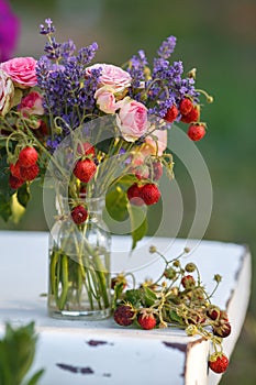 Juicy fresh bouquet of strawberries, lavender and roses stands on a white chair in a jar of water. Delicious berries and aromatic