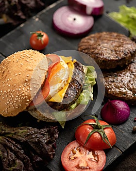 Juicy hamburger and ingredients for its preparation on black cutting board. Sesame bun with tomato, cheese, onion