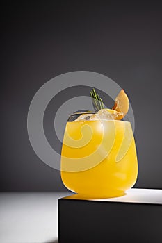 Juicy orange summer beverage with ice, green rosemary twig, peach slice in glass on pedestal shining in light on abstract modern.