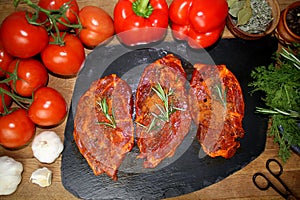 Juicy raw meat marinated with spices on black board, steaks with rosemary, red tomatoes, paprika, garlic, dill, concept of protein