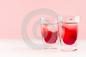 Juicy red strawberry tropical cold cocktails with ice cubes in two wet shot glasses on white wood board and light pastel pink.