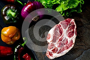 Juicy steak lies on a wild stone next to red and green chili peppers, fresh blue onions and a fresh green salad on a black wooden