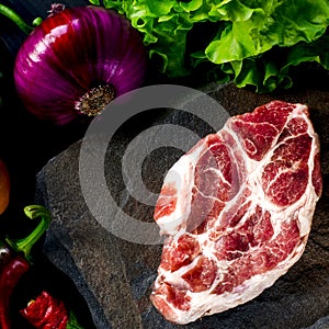 Juicy steak lies on a wild stone next to red and green chili peppers, fresh blue onions and a fresh green salad on a black wooden