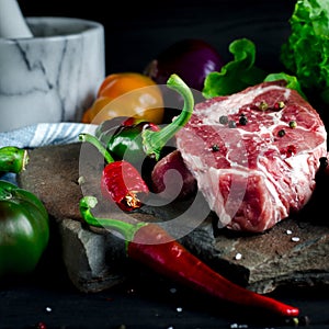 Juicy steak lies on a wild stone next to red and green chili peppers, fresh blue onions and a fresh green salad on a black wooden