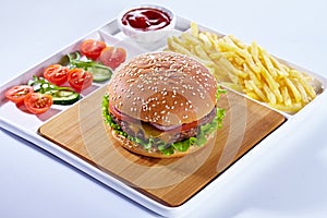 Juicy tasty hamburger on a wooden cutting board with french fried fries, vegetables and ketchup. Isolated composition on