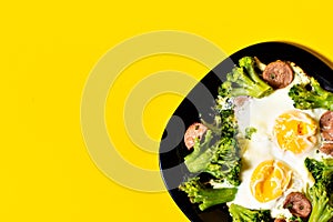 Juicy tasty healthy breakfast: fried eggs with broccoli cabbage and sausages on a black glossy plate on a yellow