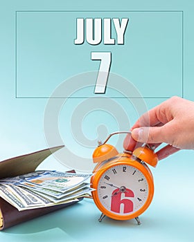 July 7th. Hand holding an orange alarm clock, a wallet with cash and a calendar date. Day 7 of month.