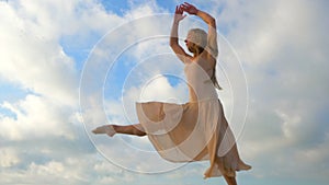 Jumping ballerina in beige silk dress and pointe on sky background. Silhouette of young beautiful blonde woman with long