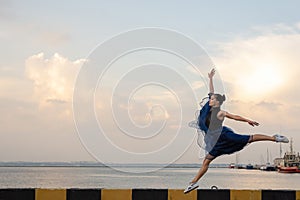 Jumping ballerina in blue dress and pointe on embankment above sea beach at sunrise.