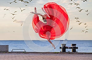 Jumping ballerina in a red flying skirt and leotard on ocean embankment or sea beach surrounded by seagulls in sky