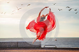 Jumping ballerina in a red flying skirt and leotard on ocean embankment or sea beach surrounded by seagulls in sky