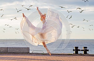Jumping ballerina in a white flying skirt and leotard on ocean embankment or sea beach surrounded by seagulls in the sky