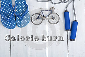 Jumping/skipping rope with blue handles, flip flops, model of bicycle on white wooden background with text calorie burn