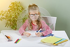 Junior schoolgirl with glasses writes something with her left hand in the notebook and sits at the table. Back to school concept.