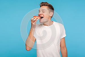 Junk food. Happy delighted man in casual white t-shirt eating donut with expression of big pleasure