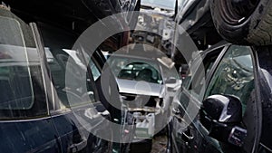 In a junkyard, a Grey car is stacked on another red car, with tires, wheels, and automotive lighting scattered around.