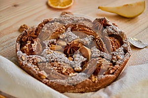 Just backed powdered pear pie decorated with white cloth, dried orange, walnuts, anise stars and spoon on the wooden board