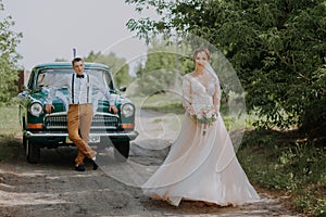 Just married wedding couple is standing near the retro vintage car in the park. Summer sunny day in forest. bride in