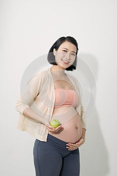 Just one apple a day. Beautiful young pregnant woman with smile and holding green apple while standing at white background