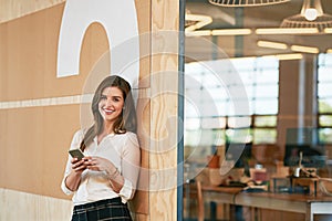 Just syncing my contacts. a smiling young woman using her cellphone while standing in an office.