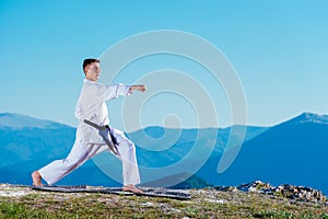 Karate man in a kimono performs a front hand kick Choku-zuki while standing on the green grass on top of a mountain