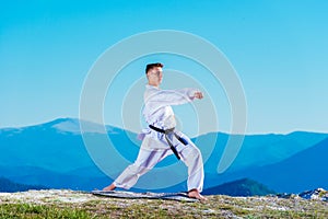 Karate man in a kimono performs a front hand kick Choku-zuki while standing on the green grass on top of a mountain