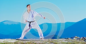 Karate man in a kimono performs a front hand kick Choku-zuki while standing on the green grass on top of a mountain