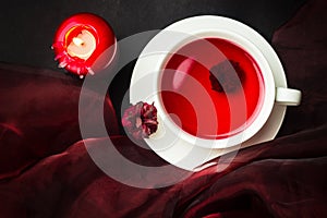 Karkade tea and a candle with dark red cloth on the background top view. The cup of red hibiscus tea and dried calyces. Herbal tea