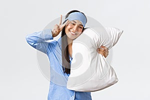 Kawaii happy smiling asian girl in blue sleeping mask and pyjama, hugging pillow and showing peace sign, feeling well