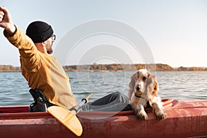 Kayaking with dogs: man stretches sitting in a row boat on the lake next to his spaniel.