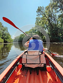 Kayaking A teenage boy in a life jacket is canoeing down the river. Trees grow along the banks. Rear view. Sports tourism and