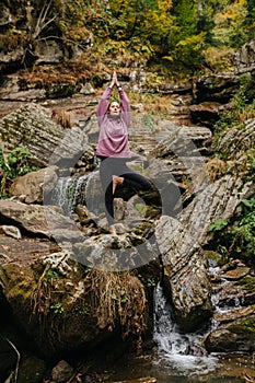 Keen woman staing on one leg on big stone, doing yoga over mountain spring
