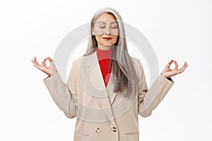 Keep calm. Asian senior woman in business suit, meditating, deep breath, inhale air and relaxing, standing over white