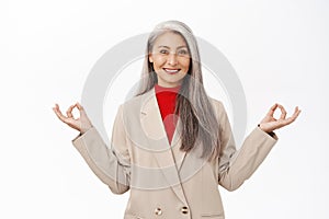 Keep calm. Asian senior woman in business suit, meditating, deep breath, inhale air and relaxing, standing over white