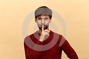 Keep silence. Photo of young man with beard wearing sweater, keeps fore finger over lips, makes silence gesture, posing against