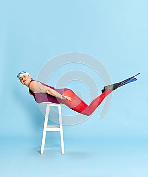 Keeping balance. Full-length image of woman in swimming suit and red tights posing with flippers on chair against blue