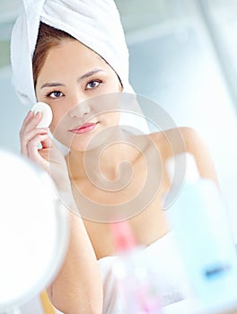 Keeping her skin soft and smooth. An attactive young Asian woman applying moisturizer with a towel on her head.